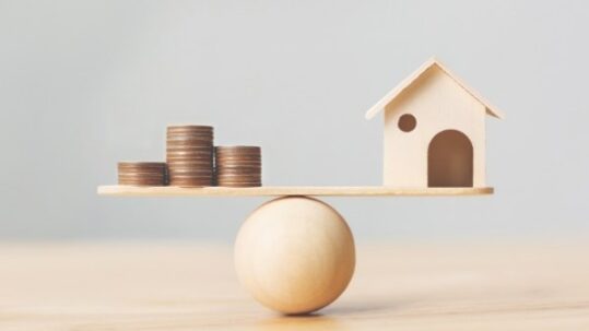 Wooden house model and money coins stack on wood scale